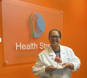 Image of a woman holding a plastic heart infront of the Health Street CPRologist sign in Houston, Texas for CPR classes offered in spanish. 